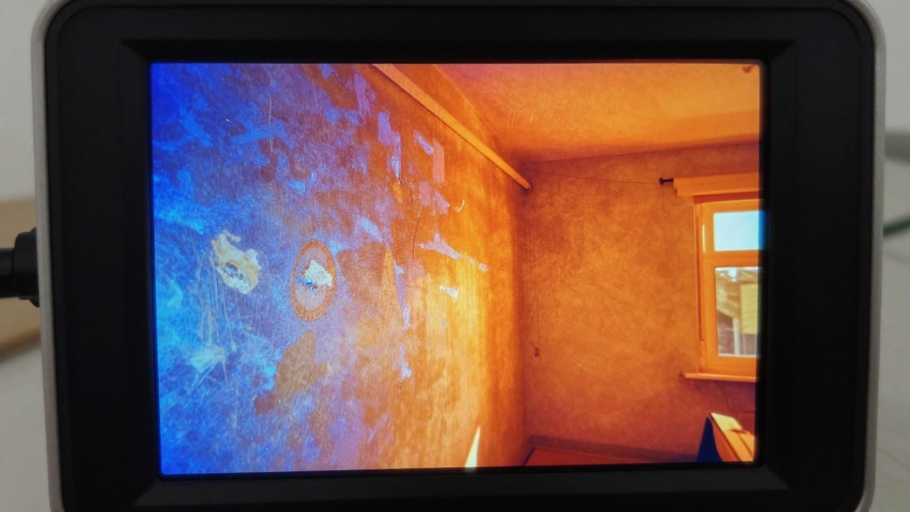 Close-up of a thermal imaging camera screen showing a house wall with cold blue patches and warm orange areas in a residential room